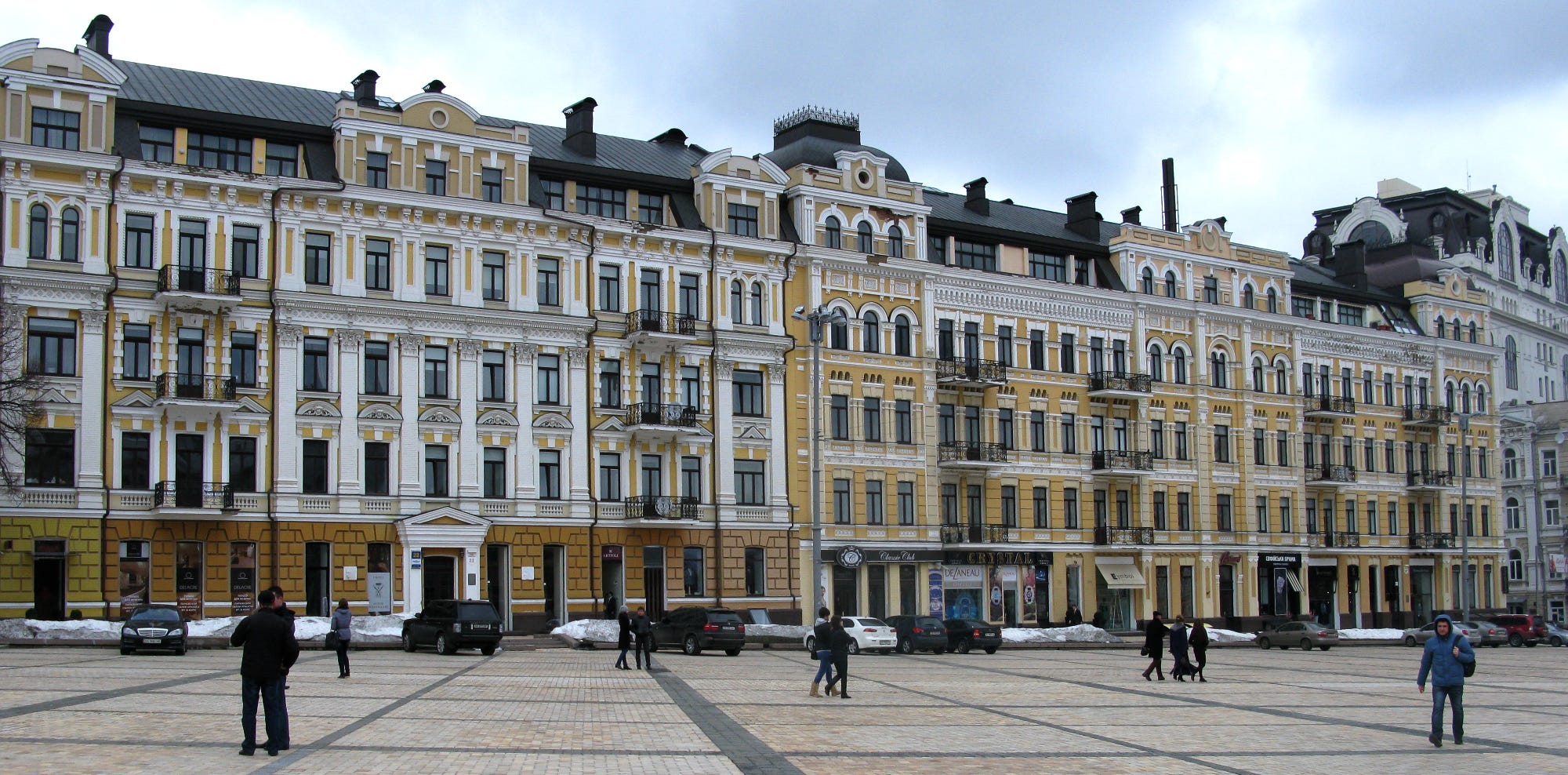 A row of attached buildings in an ornate style reminiscent of Parisian style Haussmann architecture, with a broad plaza in the foreground and nearly a small figures walking around in winter clothes.