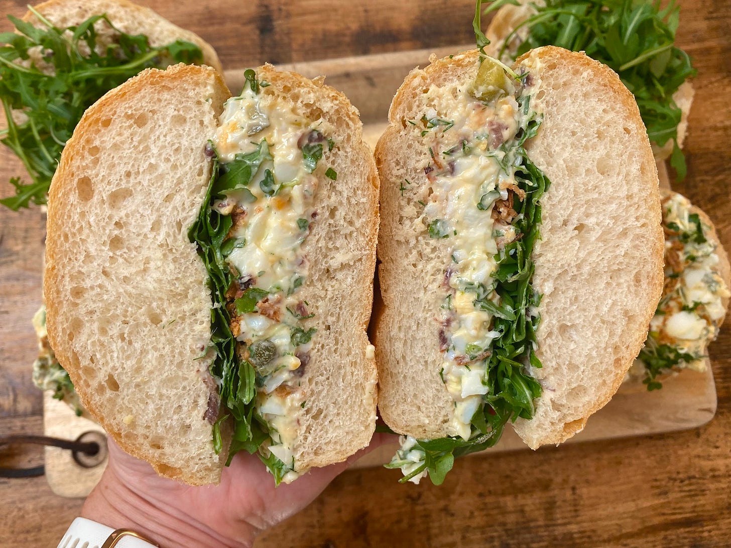 A cut-open cross section of a sandwich filled with egg mayonnaise, salad leaves, on thick white bread. Behind the sandwich, there are other sandwiches on a chopping board on a table.
