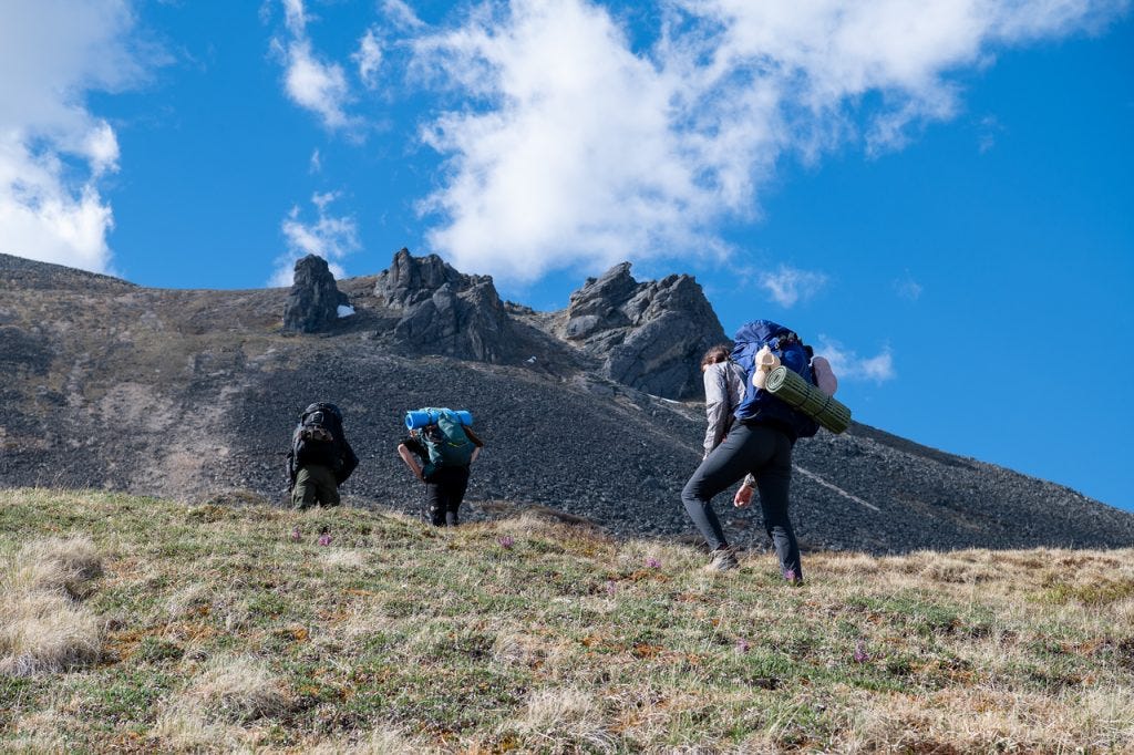 Seen from below, three hikers with packs climb a grassy alpine slope toward a ridge topped with jagged rock spires, against a blue sky with scattered cloud.