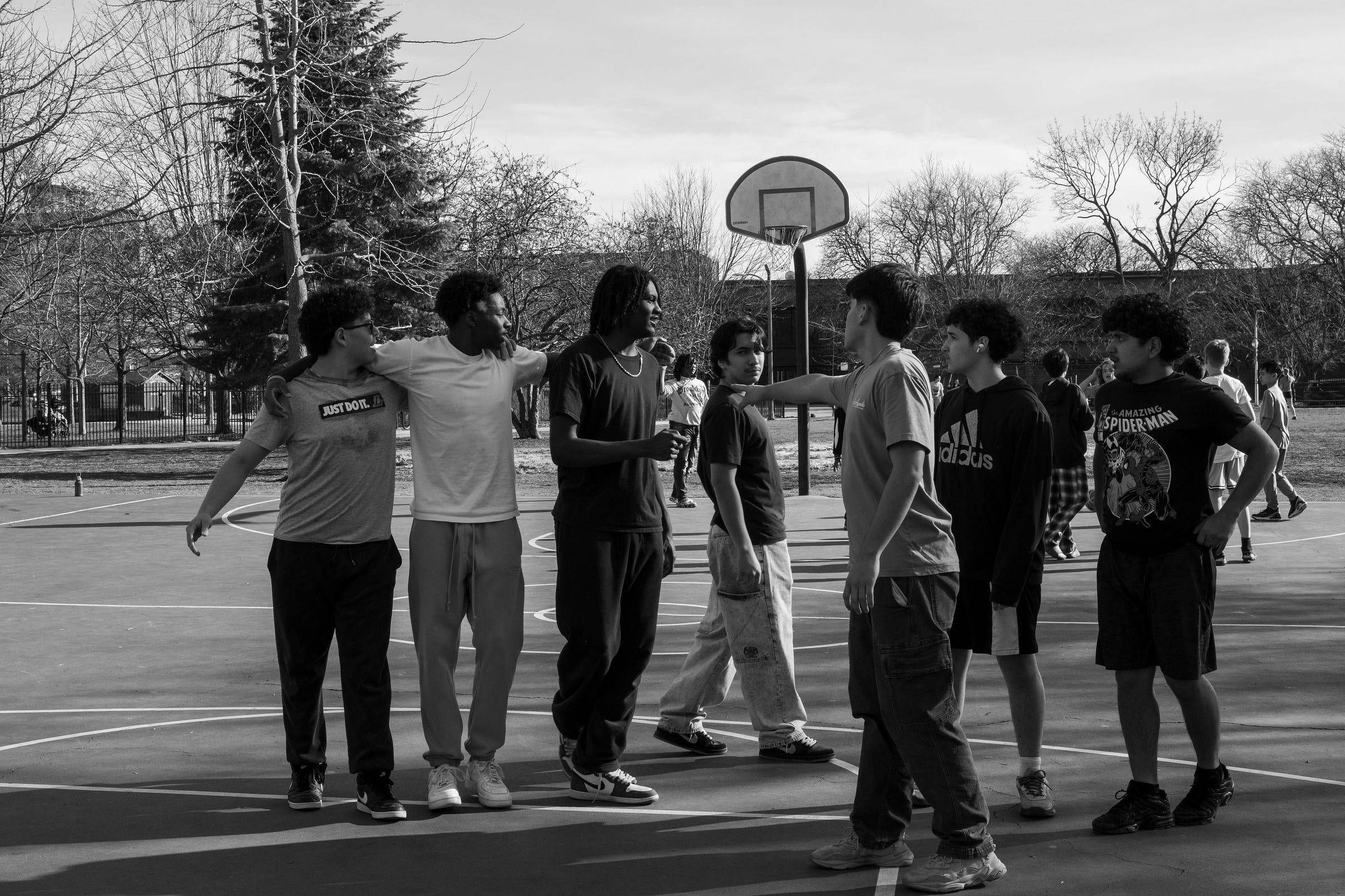 A black-and-white photograph of a group of young men gathered on an outdoor basketball court, engaged in conversation. Some have their arms around each other, while others gesture animatedly, possibly discussing the game. A basketball hoop stands in the background, along with leafless trees and a fenced park area. The scene captures camaraderie, movement, and the unspoken bonds formed through shared spaces and experiences.