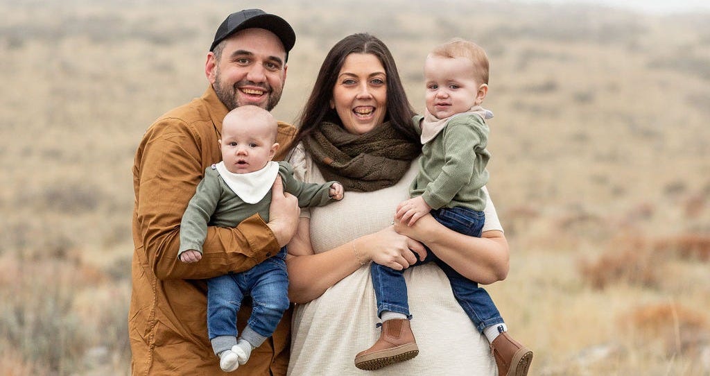 Katlyn Slone stands outdoors smiling with her husband Jared and their two young sons, Michael and Matthew. Jared holds Michael, and Katlyn holds Matthew, both toddlers dressed in green tops and jeans. The family is bundled warmly against a misty hillside background. Katlyn Slone stands outdoors smiling with her husband Jared and their two young sons, Michael and Matthew. Jared holds Michael, and Katlyn holds Matthew, both toddlers dressed in green tops and jeans. The family is bundled warmly against a misty hillside background.