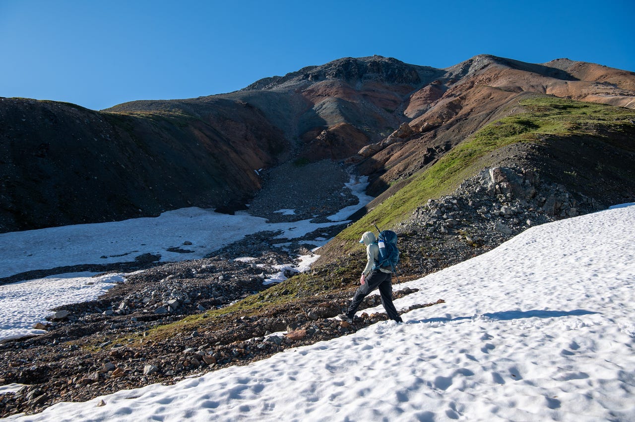 A hooded hiker in a pale sun shirt and dark pants, teal pack on, walks across a patch of old snow on a green tundra bench. Above, a mountainside striped in rust-red, ochre, and grey rises to a dark summit, a snow-filled gully threading down it under a clear blue sky.
