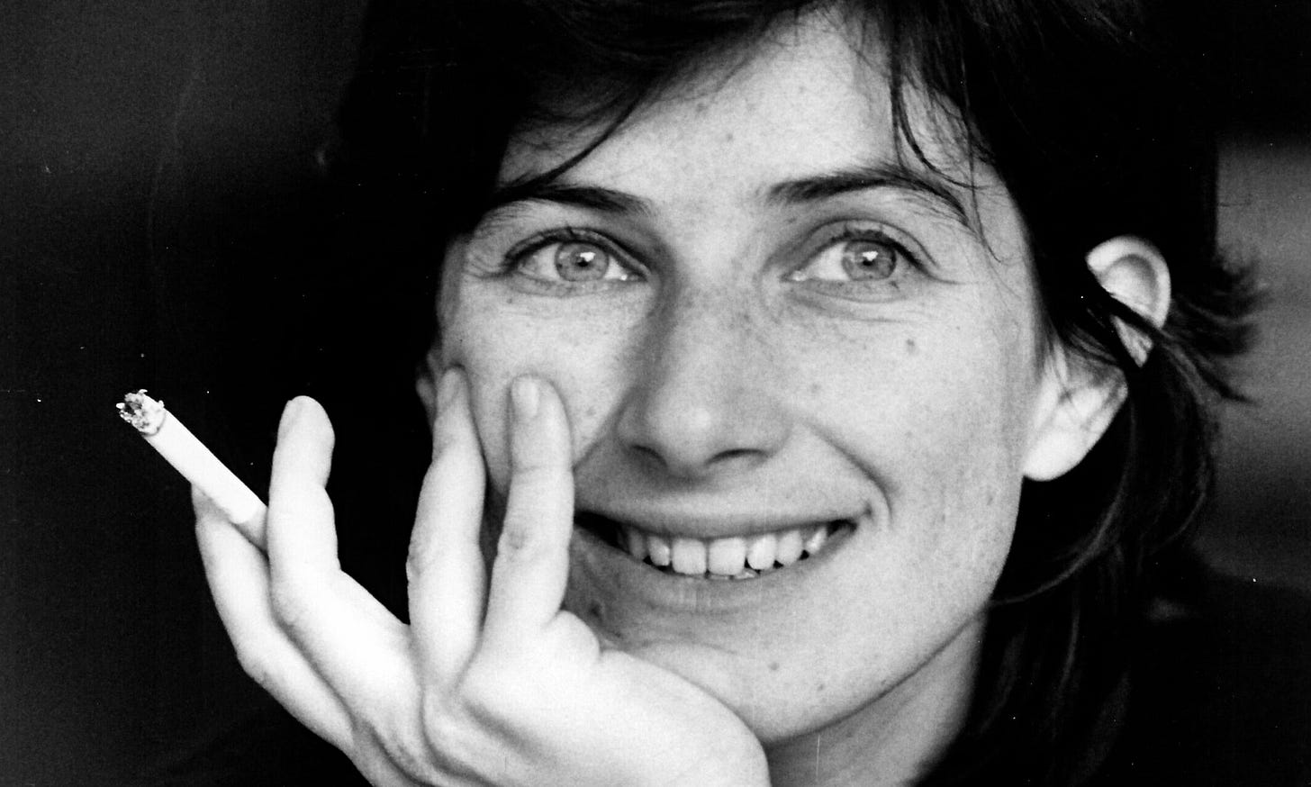 Portrait of a young Chantal Akerman, a pale-skinned woman with dark hair and light eyes, smiling and gazing out beyond the camera. Her hand pressed to her face holds a lit cigarette between index and middle fingers.
