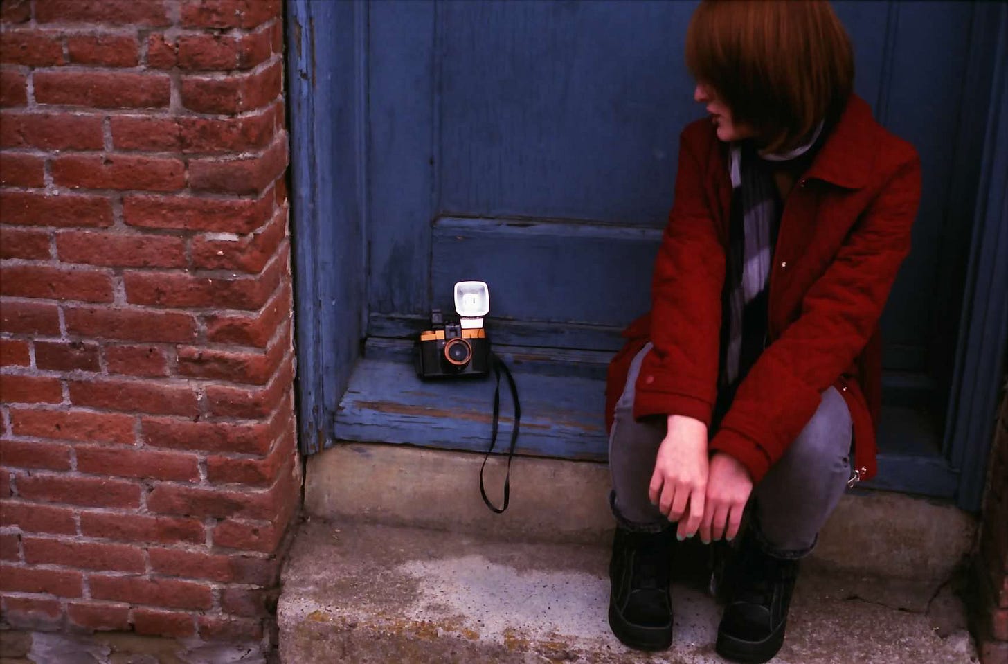 A photograph of Sarah, who sits on a stoop before a blue-lavender door and contrasting deep red brick wall. Sarah's face is somewhat hidden, as she looks toward a film camera sitting on the ground next to her. She wears a red jacket and scarf.