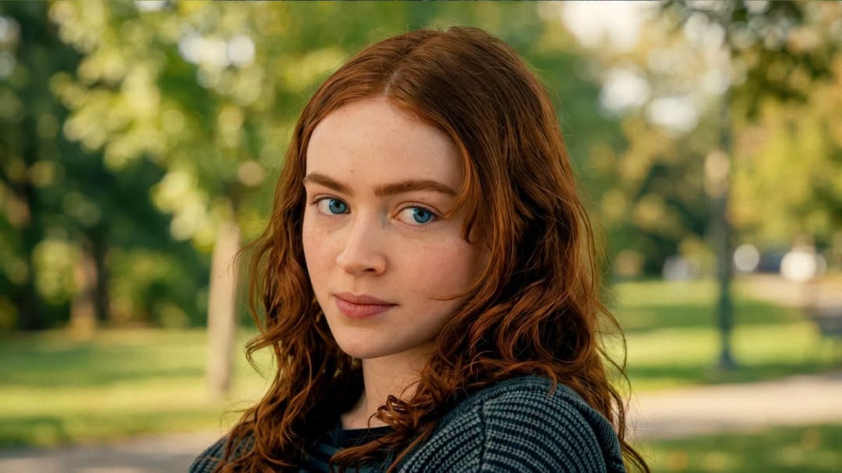 An outdoor, medium close-up shot captures actress Sadie Sink, looking over her shoulder with a focused expression against a softly blurred park background. She is depicted with her characteristic long, wavy red hair parted down the middle, with a few strands framing her fair-complexioned face. Her striking blue eyes are directed toward the camera, and her lips are closed in a neutral, calm expression. She is wearing a dark, textured grey knit cardigan or sweater over a striped shirt. The warm, natural sunlight filters through the green trees in the background, creating a soft bokeh effect and highlighting the texture of her hair and clothing.