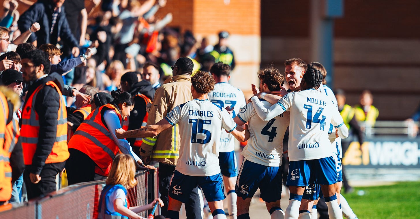 Huddersfield Town players, wearing a mint and white away strip, celebrate a goal, with fans and stewards in the background.