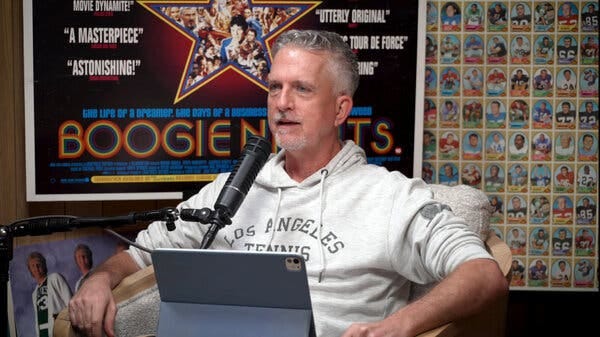 A man wearing a Los Angeles tennis sweatshirt sits in front of a microphone, with a poster for “Boogie Nights” behind him.