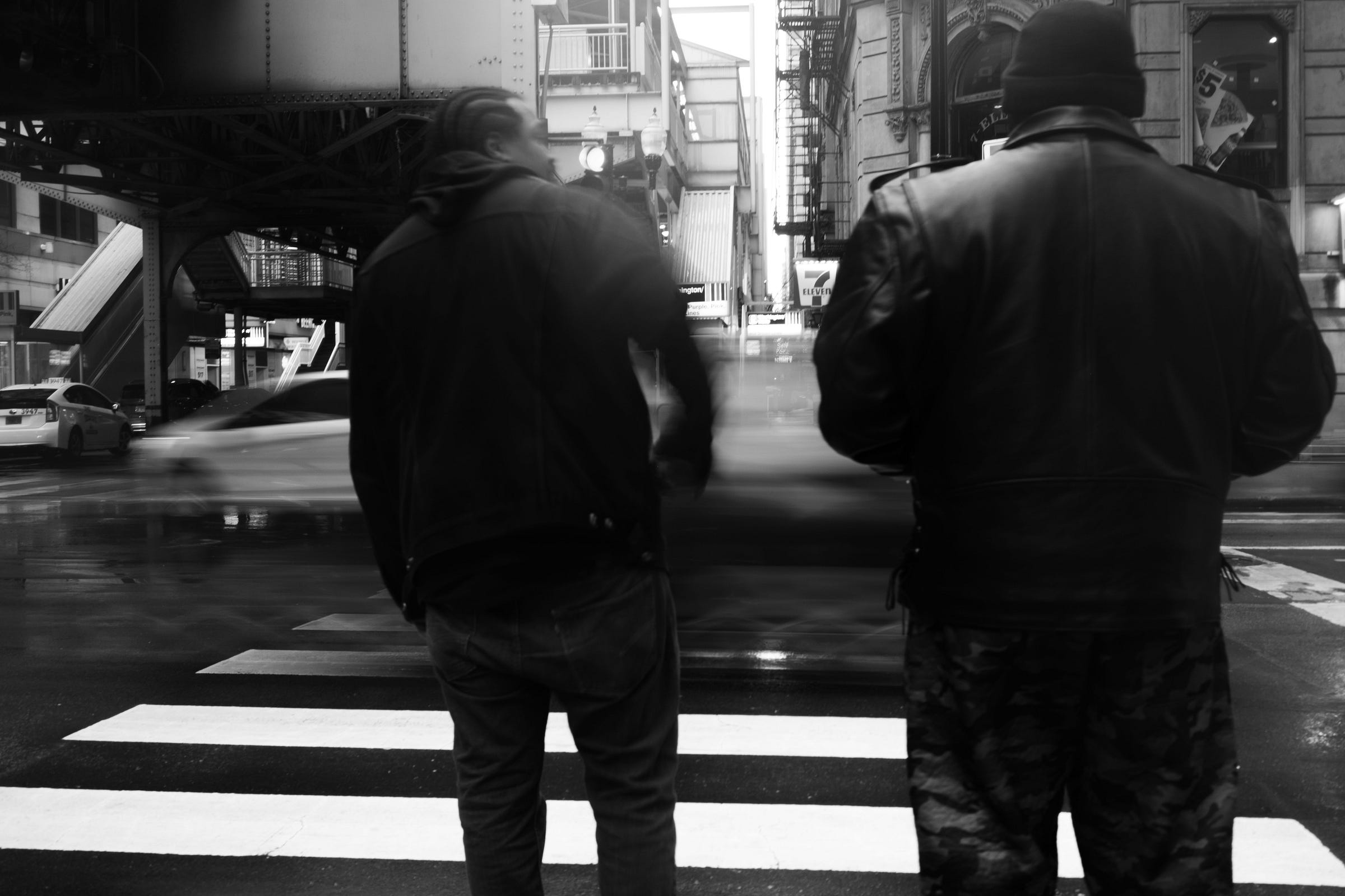 Two men in leather jackets stand at a crosswalk under an elevated train, watching as a blurred car speeds past on the rain-slicked street. Two men in leather jackets stand at a crosswalk under an elevated train, watching as a blurred car speeds past on the rain-slicked street.