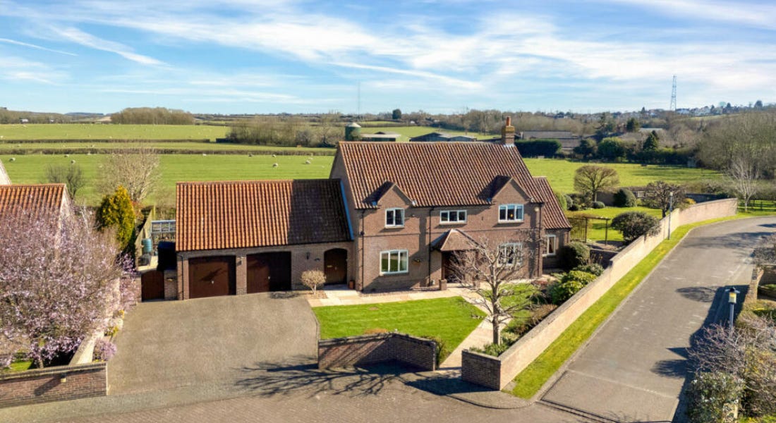 A large brown house with a garage to the left of it, a lawn with sheep on behind it, and a driveway and road in front of it