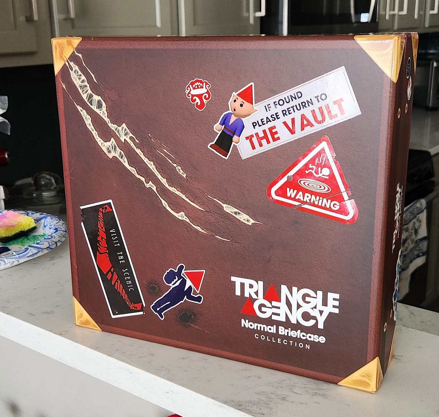 A photo of the Triangle Agency Normal Briefcase box set sitting on a kitchen countertop. It is a box designed to look like a brown leather briefcase with gold corners, adorned with a variety of stickers and bearing a large scratch that extends from the top left to the center of the box.
