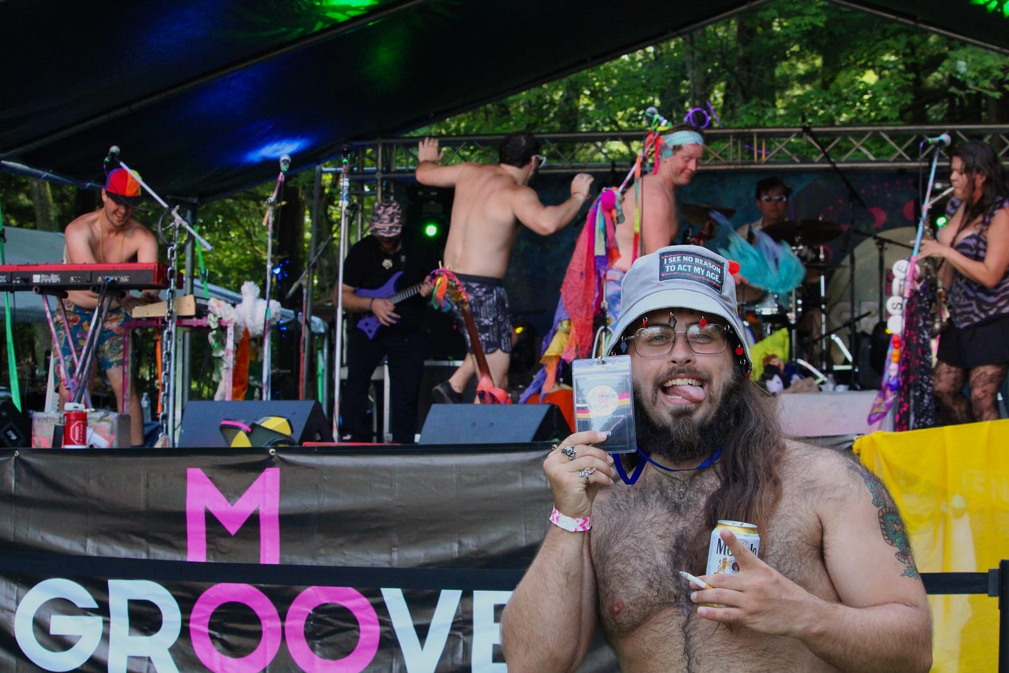 A festivalgoer holds up his MoonGroove badge in the foreground while Sneezy performs behind him on the main stage, band members dancing and playing keyboard and guitar under a tented outdoor stage. A festivalgoer holds up his MoonGroove badge in the foreground while Sneezy performs behind him on the main stage, band members dancing and playing keyboard and guitar under a tented outdoor stage.