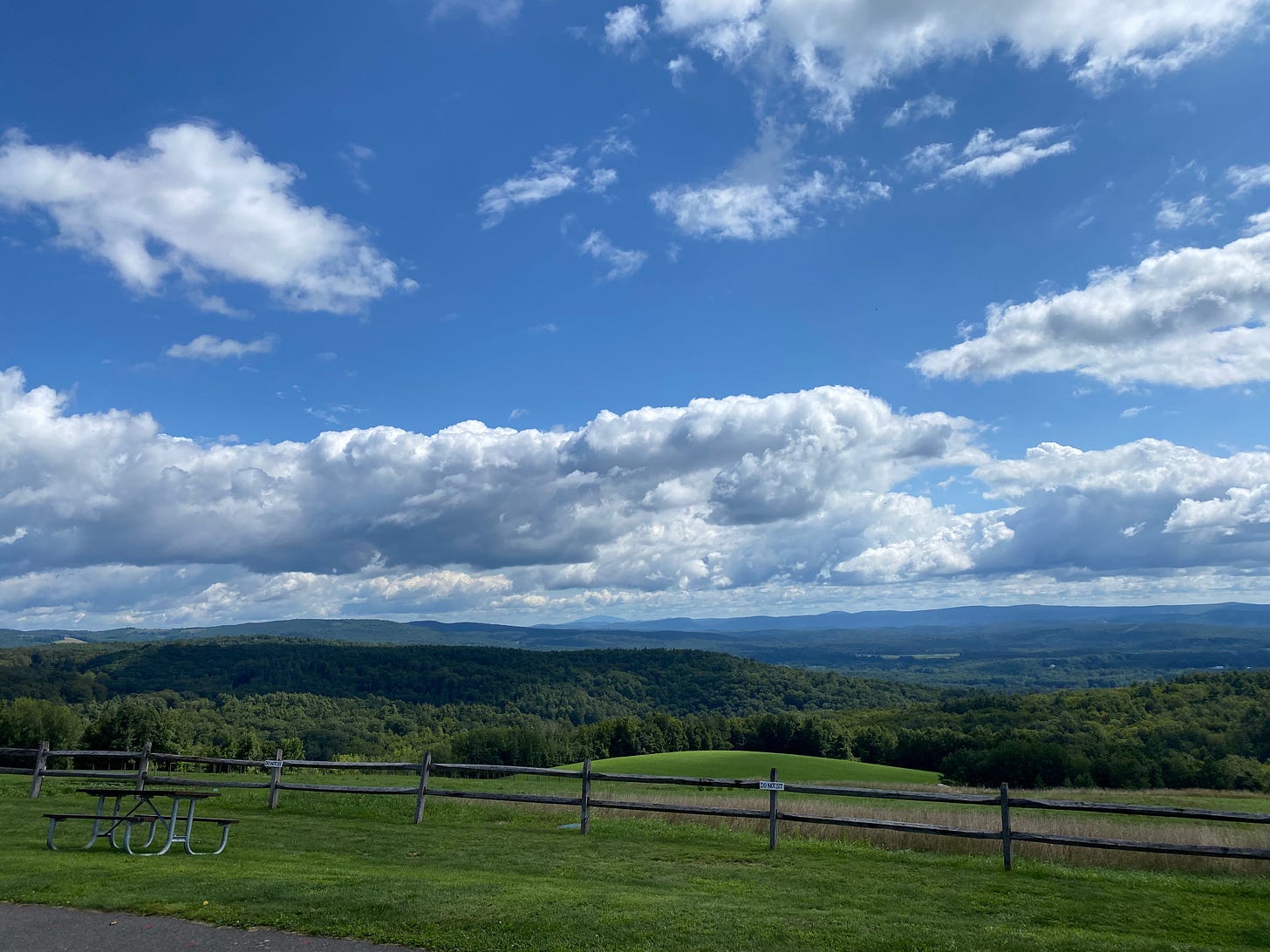 A sweeping view of the Valley from Apex Orchards, looking across green, tree-covered hills to blue mountains beyond. The sky is bright blue and full of bright white clouds.