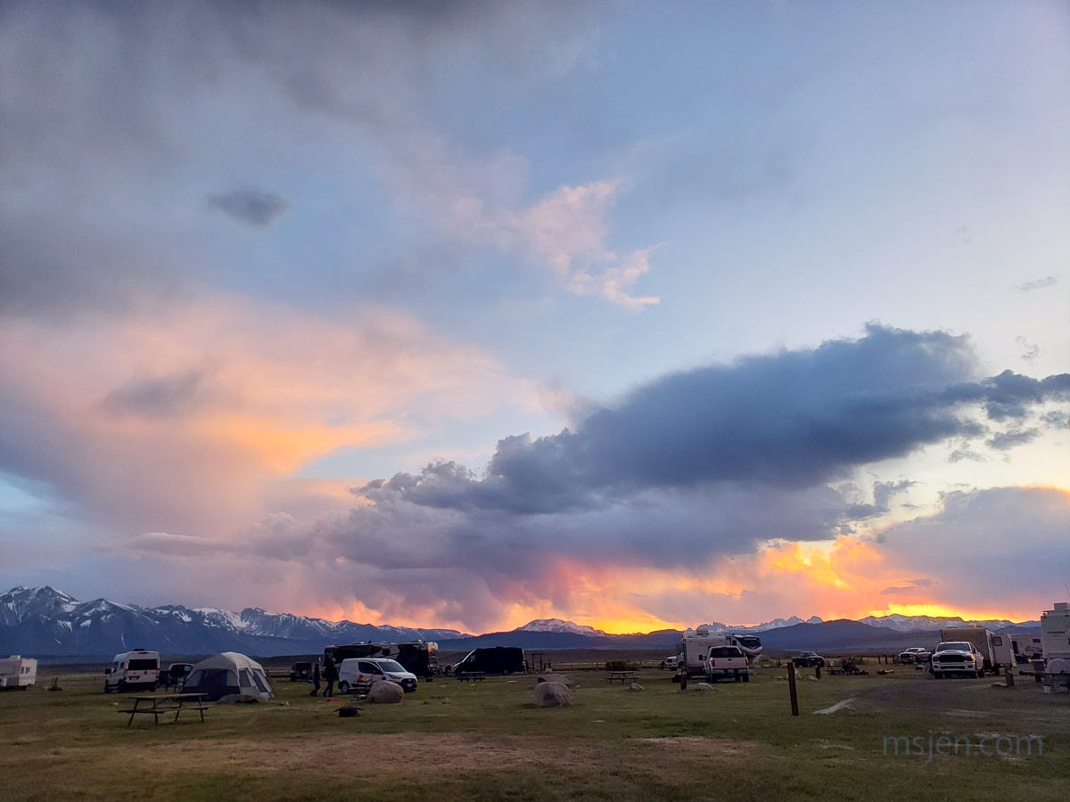 Foreground of photo is a dusky summer grasslands campgrounds full of Memorial Day Weekend campers in tents, motorhomes, and trailers. The middle ground is a backlit horizon of dark, tall mountains with snow still high up and in the canyons. The background is a bright orange sunset sky with dark clouds and at the very top of the image a blue sky. Photo by Jenifer Hanen.