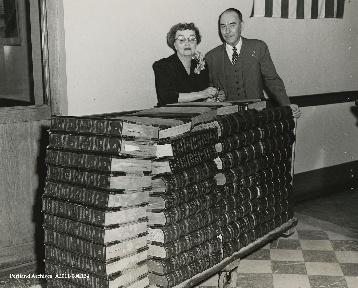 Black and photo of a woman and man behind stacks of large volumes