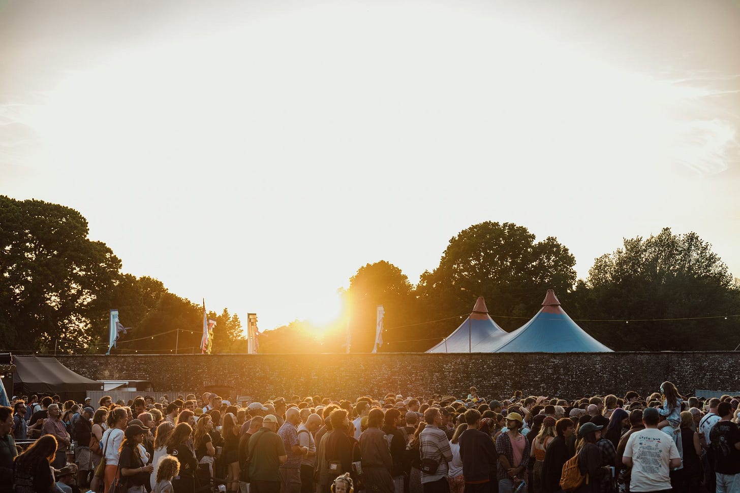 A beautiful golden-hour photograph of a large, dense festival crowd gathered at the Green Man Festival in Powys, Wales. The warm setting sun glows directly behind trees and large blue-and-white pointed festival tents, casting a bright haze over the festival-goers.