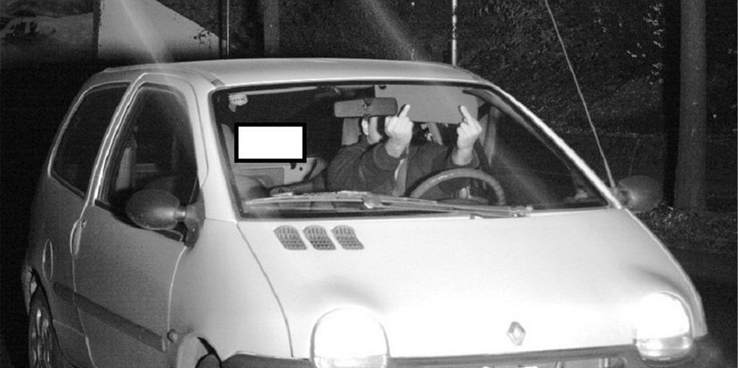 Fahrer im Kleinwagen zeigt beide Mittelfinger in Richtung Kamera, während er fährt.