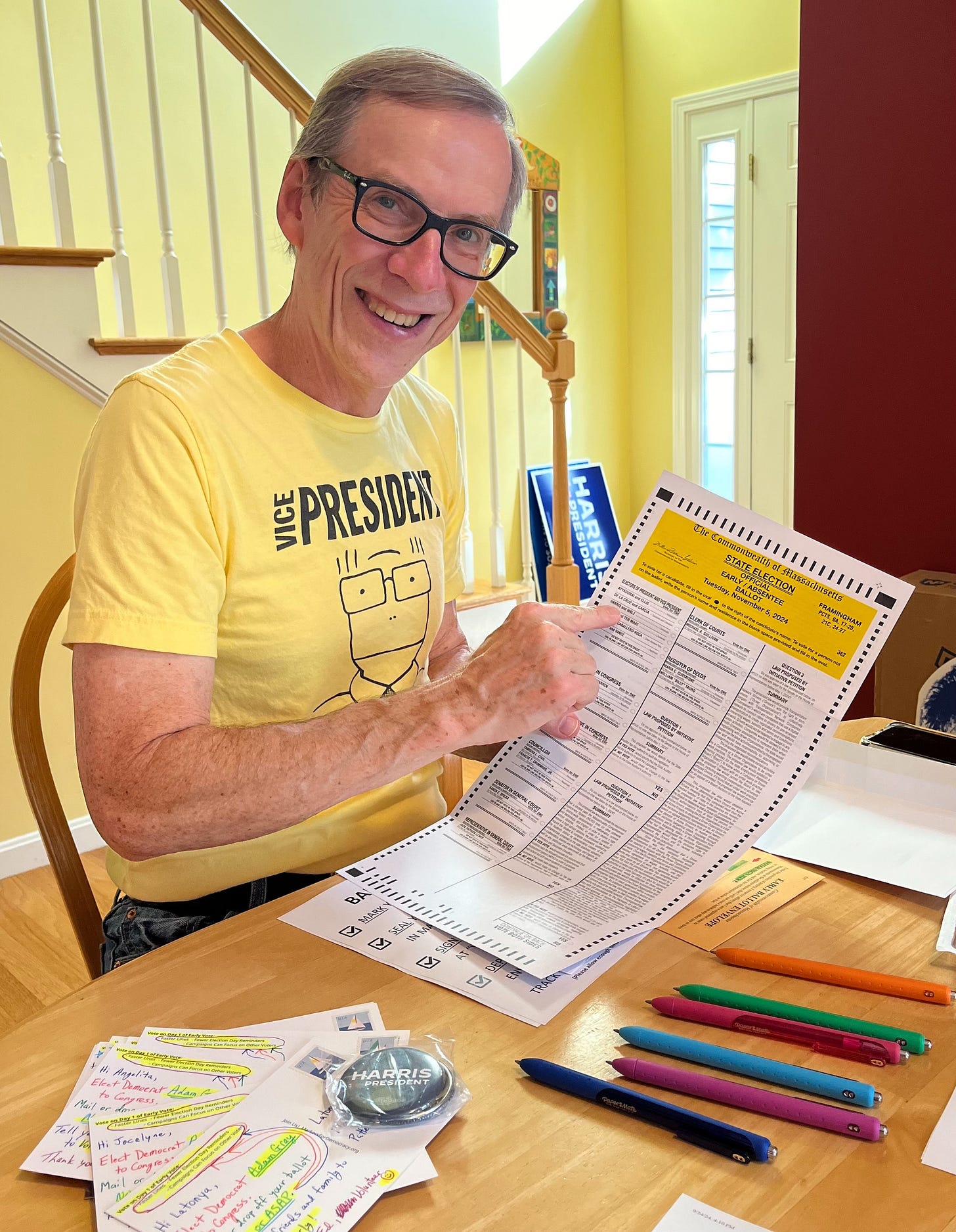 A smiling man in a yellow shirt sits at a table with his vote by mail ballot, pointing to Kamala Harris's name. There are colorfully written postcards (addresses artfully hidden by a Harris for President button) and an array of colored pens on the table. In the background is the Harris for President yard sign that Suz got before she got the one that says Harris/Walz that's in her yard right now.