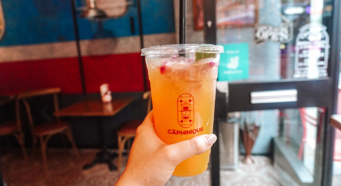 A hand holding an iced orange drink in a plastic cup, with a restaurant behind it