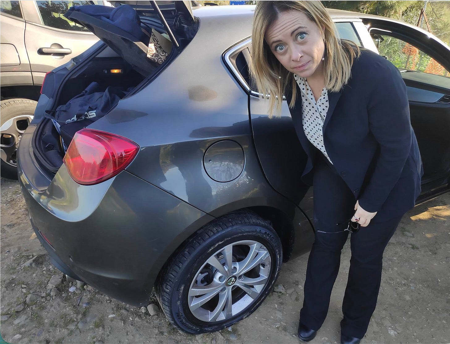 Giorgia Meloni accanto a un’auto con una ruota posteriore sgonfia.
