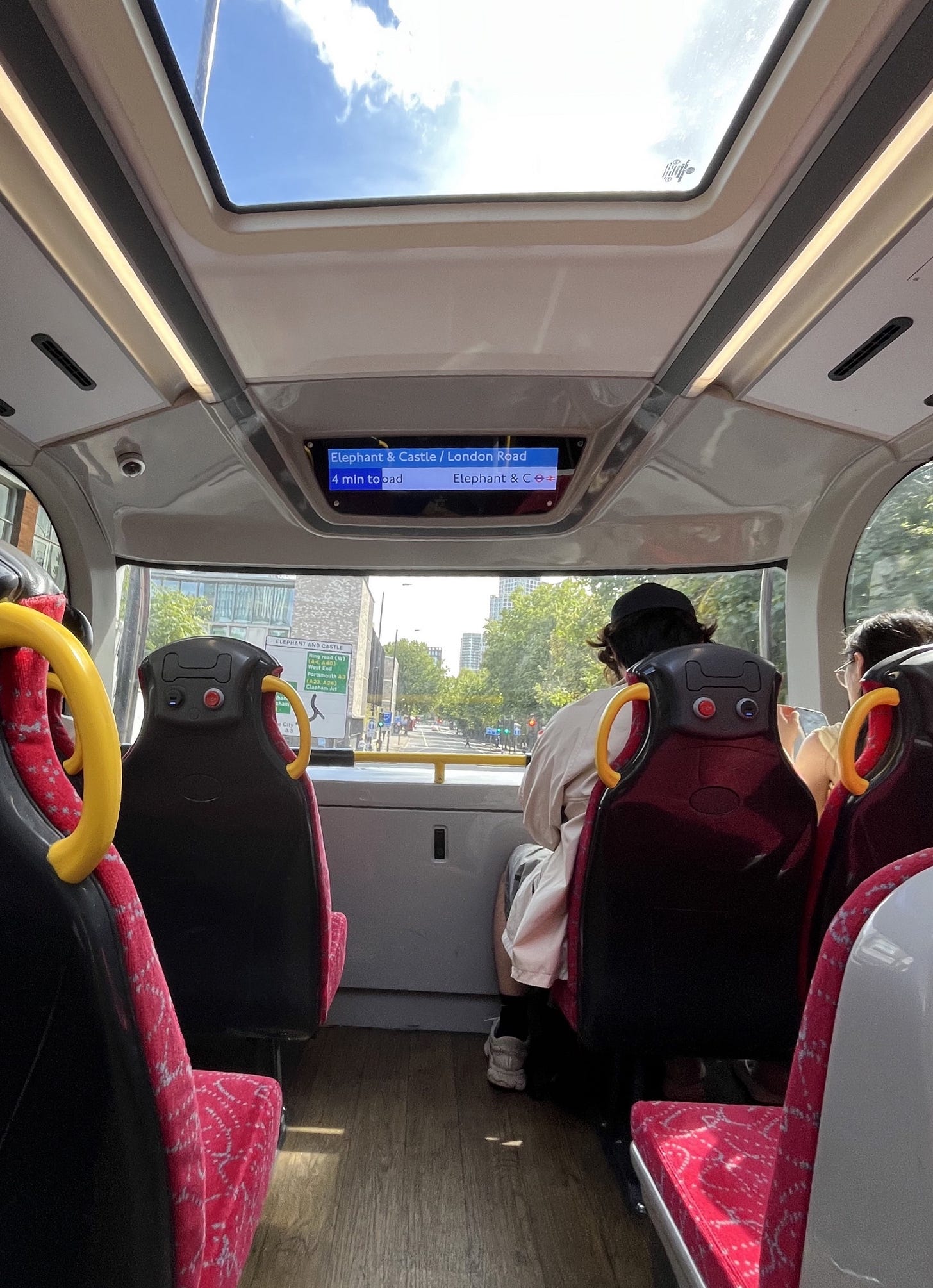 Photo of the front upper deck of a bus on the 63, showing colour screen, high back seats with charging and stop buttons, wooden flooring and skylight.