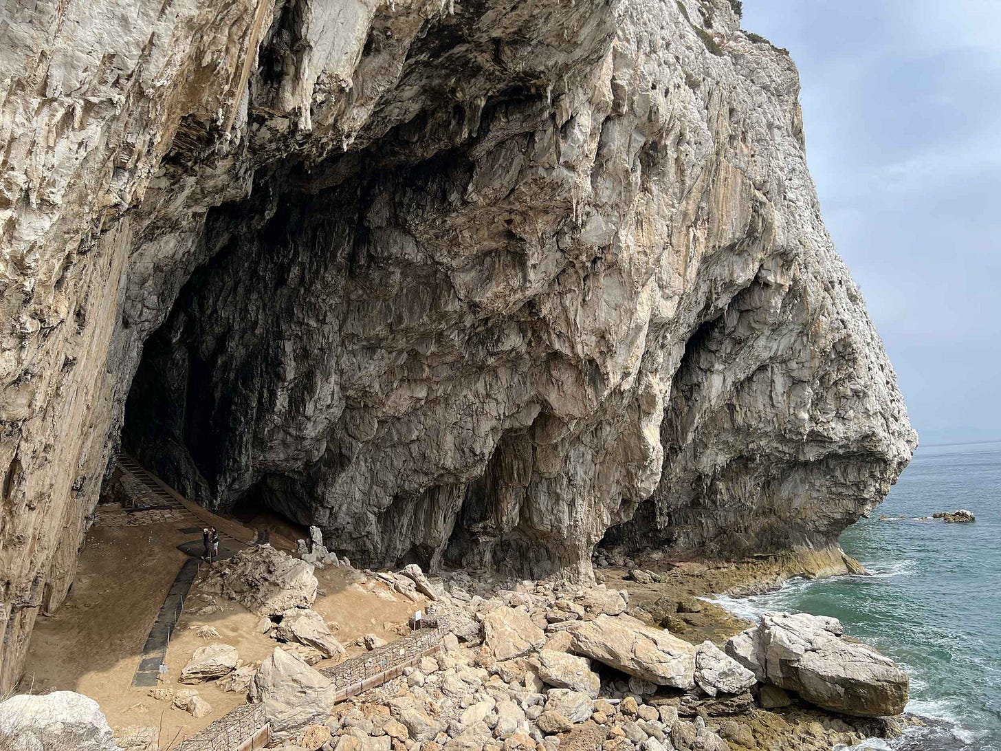 A large cave mouth with people standing beneath for scale, the rocky beach and sea to right of frame A large cave mouth with people standing beneath for scale, the rocky beach and sea to right of frame