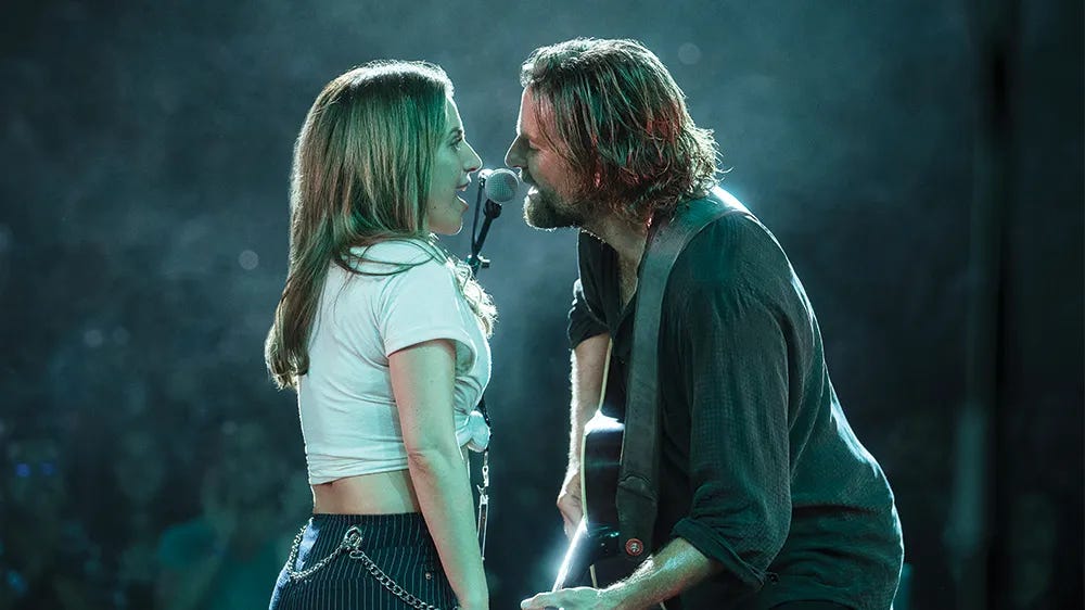 Screenshot from the film 'A Star is Born.' In this scene, two musicians share a microphone on stage in an intimate performance moment. A woman in a white crop top and striped pants stands face-to-face with a bearded man in dark clothing who is playing guitar. They're singing into the same microphone against a smoky, blue-green lit background that creates a moody concert atmosphere.