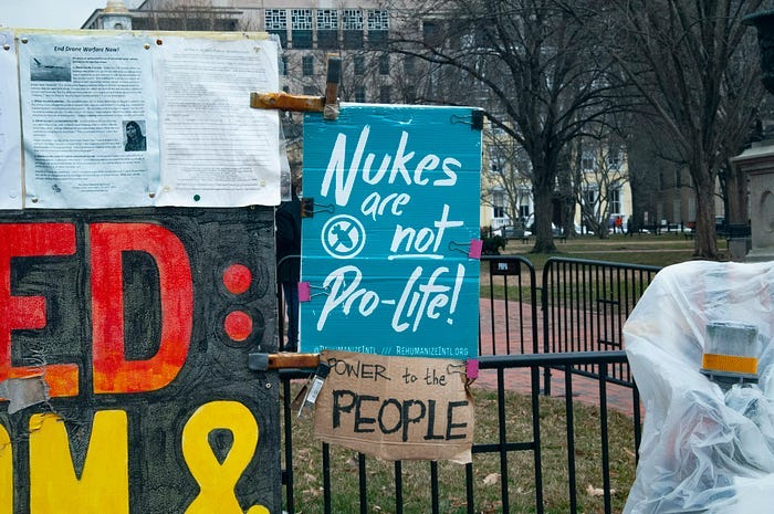 A painted sign hanging from a wall. It is pale blue with white lettering, and it reads: “Nukes are not pro-life!” A painted sign hanging from a wall. It is pale blue with white lettering, and it reads: “Nukes are not pro-life!”