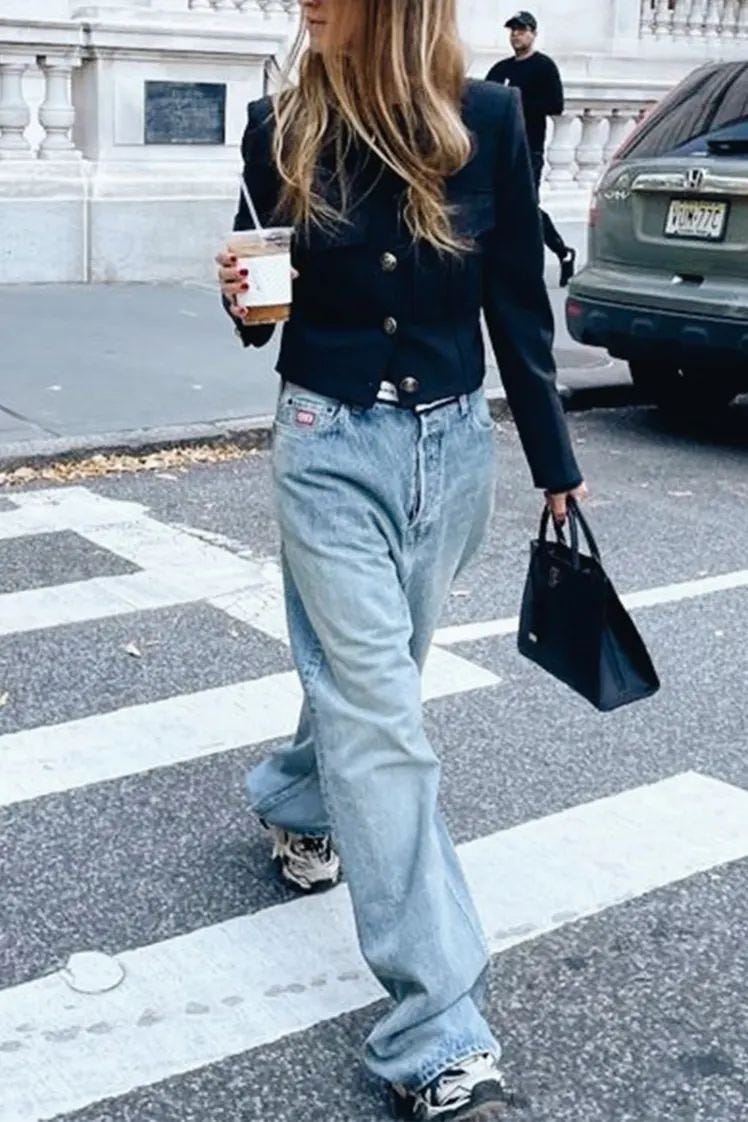 Mujer con look sencillo y atemporal (jeans, zapatillas, rebeca y café en la mano) cruzando un paso de cebra representando la moda real y el estilo honesto