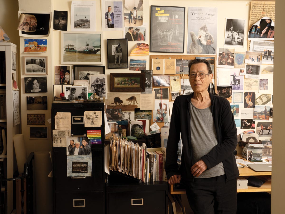An elderly white woman stands next to filing cabinets. Numerous pieces of paper―posters, photos of dancers, newspaper clippings―are pinned up behind her.