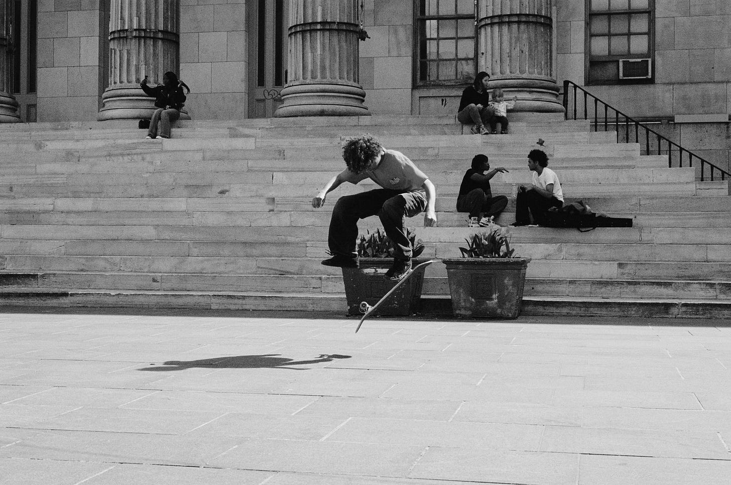 A teenage boy skateboarding is suspended in the air while doing a trick in a public plaza. On the steps behind him sits a girl taking a selfie on her phone, a woman with a young child, and two teenage boys having a conversation while sitting next to their skateboards.