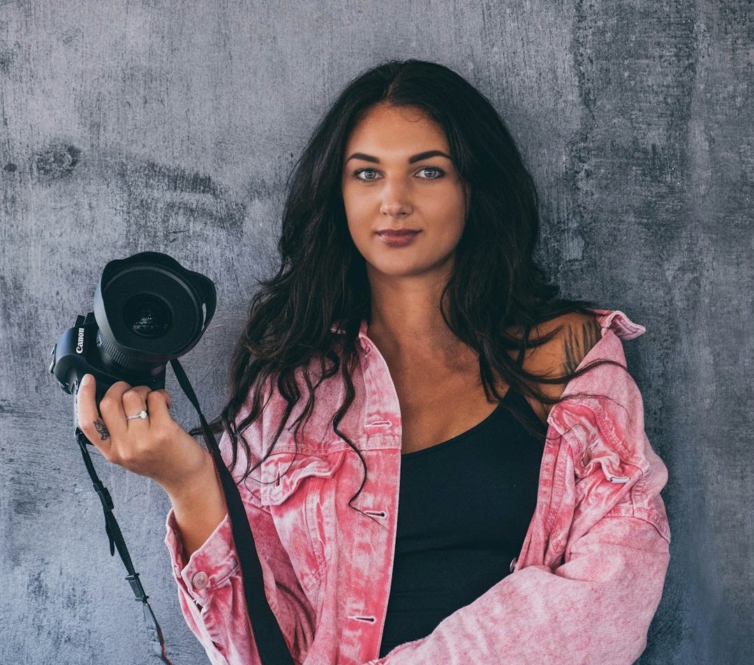 woman standing in front of gray wall holding black DSLR camera woman standing in front of gray wall holding black DSLR camera