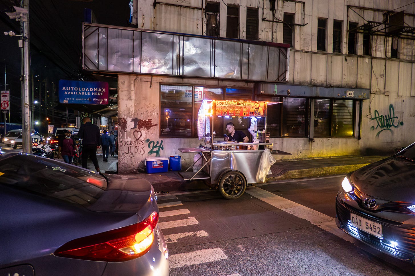 Nighttime street scene. Cars stop at an intersection while a street vendor sits with cart parked up against the curb. The vendor is not busy - he is looking at his phone. His cart is in front of an empty corner store whose sign that wraps around the front and side has been removed, leaving behind a bare, battered, metal frame. There is grafitti on the dirty white walls. Two blue coolers sit by the building, presumably part of the street vendor's stuff.