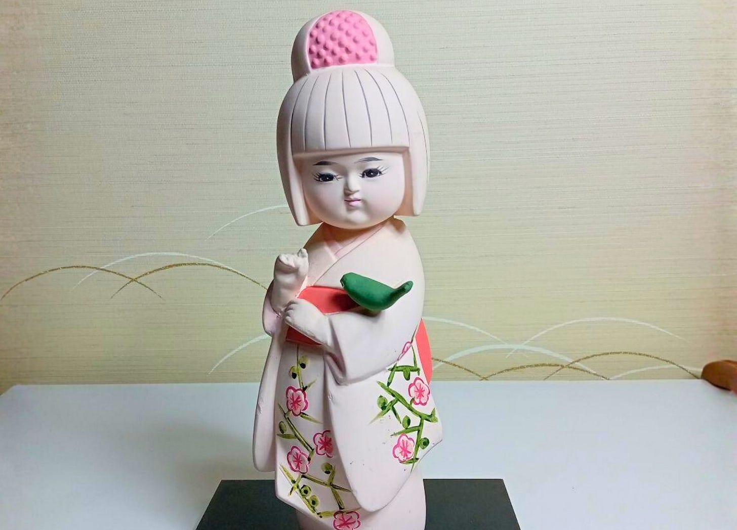A traditional Japanese ceramic doll of a young girl in a pink kimono decorated with plum blossoms, holding a green bird on her arm.