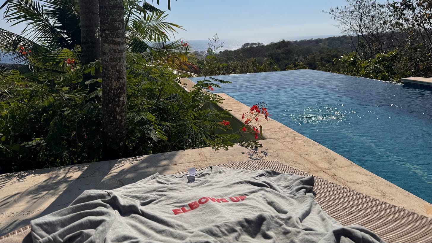 A grey "Elbows Up" t-shirt on a sun lounger in front of a pool atop a jungle hill in Costa Rica. A grey "Elbows Up" t-shirt on a sun lounger in front of a pool atop a jungle hill in Costa Rica.
