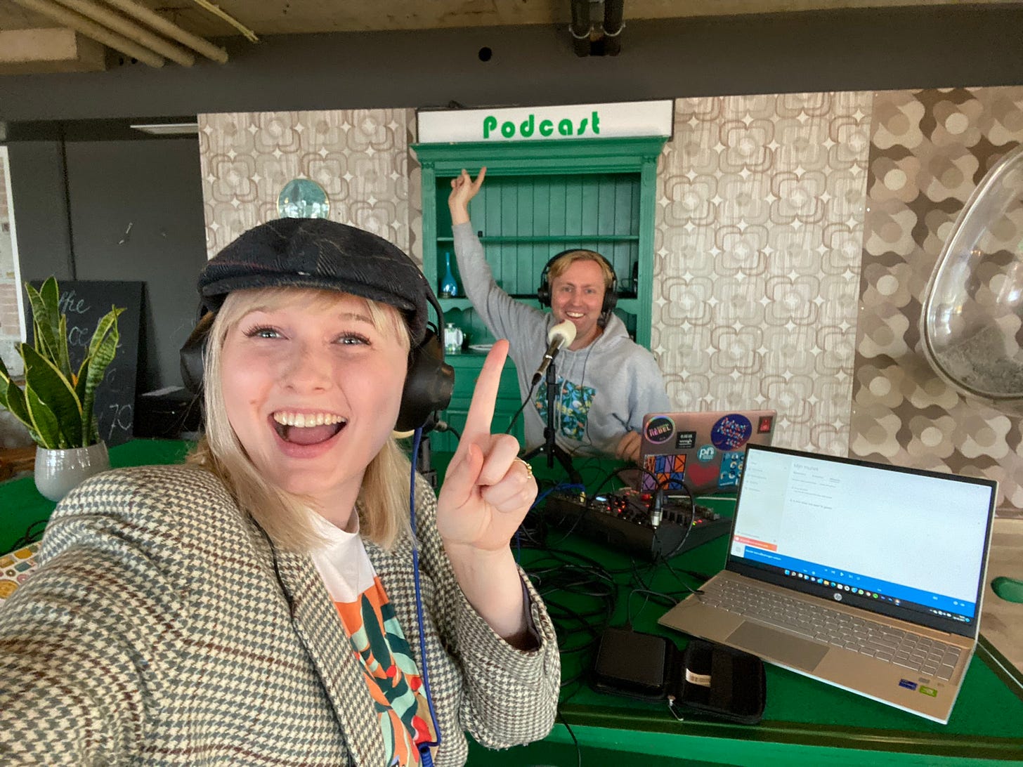 foto van Lieven en Femke die in de Podcastruimte van de Toluistuin aan een groene tafel zitten. Er staan laptops en audio spullen op tafel, ze lachen heel blij