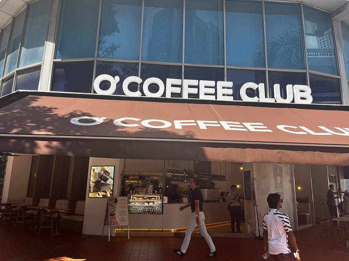 Left photo: exterior of the O'Coffee Club shop. A brown awning hangs above the shop's entrance and O'Coffee Club is written in white letters on the awning. Right photo: A slice of cheesecake, strawberry lemon soda and a bottle of water on a cafe table. 