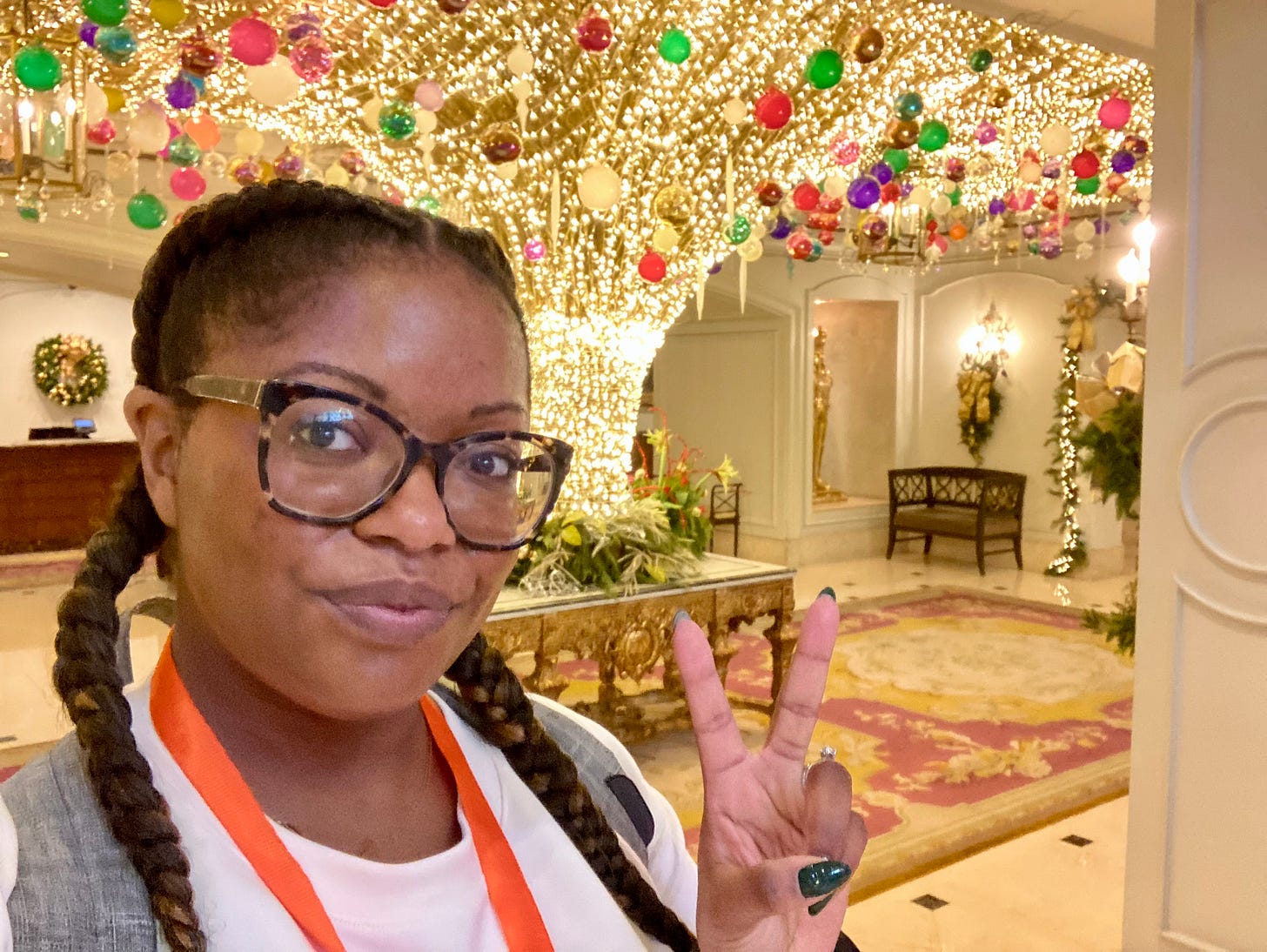 Picture of a Black woman with glasses and French braids holding up the peace sign in the lobby of the Ritz-Carlton in New Orleans. There are Christmas lights and ornaments on the ceiling behind her.