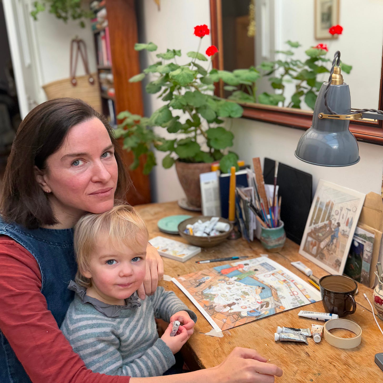Anna Neima sitting at a wooden desk with a young child on her lap, working on an illustration, with paints, sketchbooks, and a desk lamp nearby.