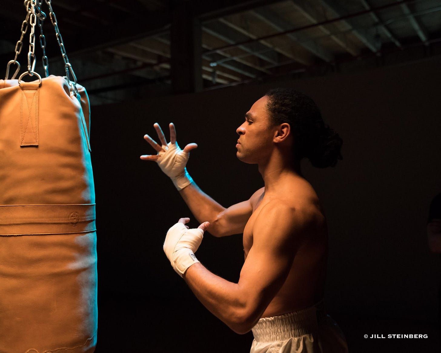 In a photo by Jill Steinberg, Brandon Kazen-Maddox, a shirtless black masculine person, is facing a punching back with a grimace taped hands.