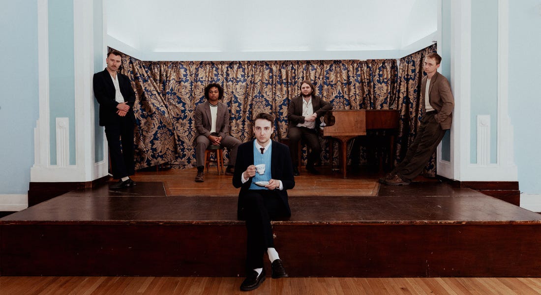 Five men from the band Victory Lap sat on top of a dark brown wooden stage with a blue wall behind them