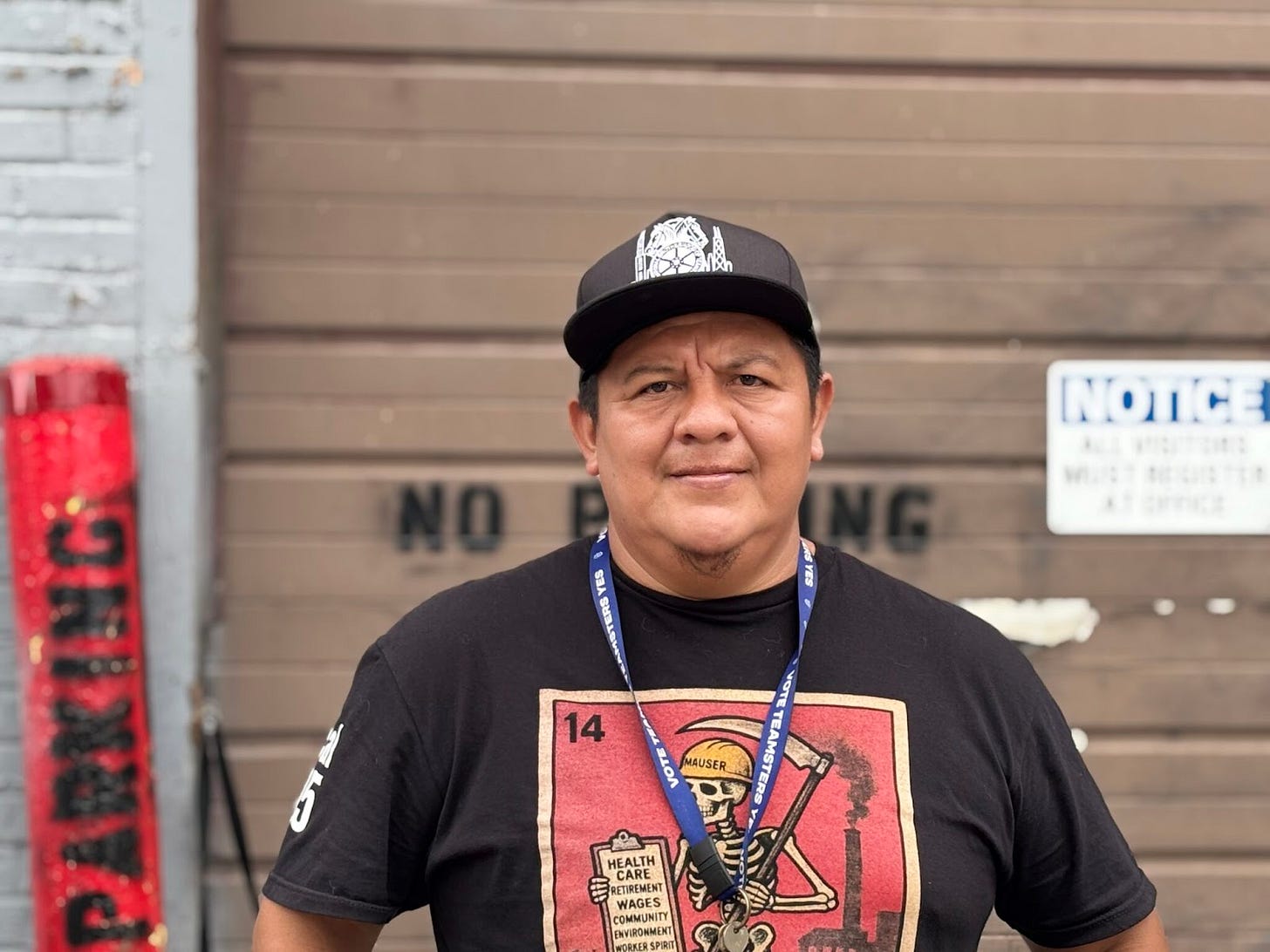 a Brown skinned man wearing a black teamsters cap and a black tee shirt with a skeleton wearing a yellow construction hat stands in front of a garage door a Brown skinned man wearing a black teamsters cap and a black tee shirt with a skeleton wearing a yellow construction hat stands in front of a garage door