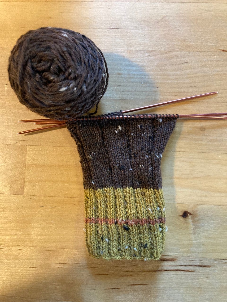 The beginning of a handknit sock in mustard yellow, orange, and brown tweed yarn. The sock is on knitting needles and next to a ball of yarn.