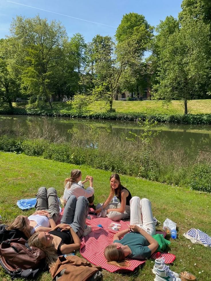 This may contain: group of people laying on the grass next to a river and picnicking in the park