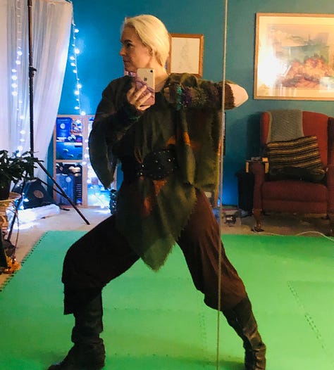 Two mirror-selfies on the green screen floor: a drab brown and olive ratty hunting outfit and a pretty-in-pink-and-sand pants and tunic outfit. And a pro shot of my She-Wolf fur and feathers wild-woman outfit in a dynamic, claw-handed pose with leg poised to kick and gaze aimed and fixed.