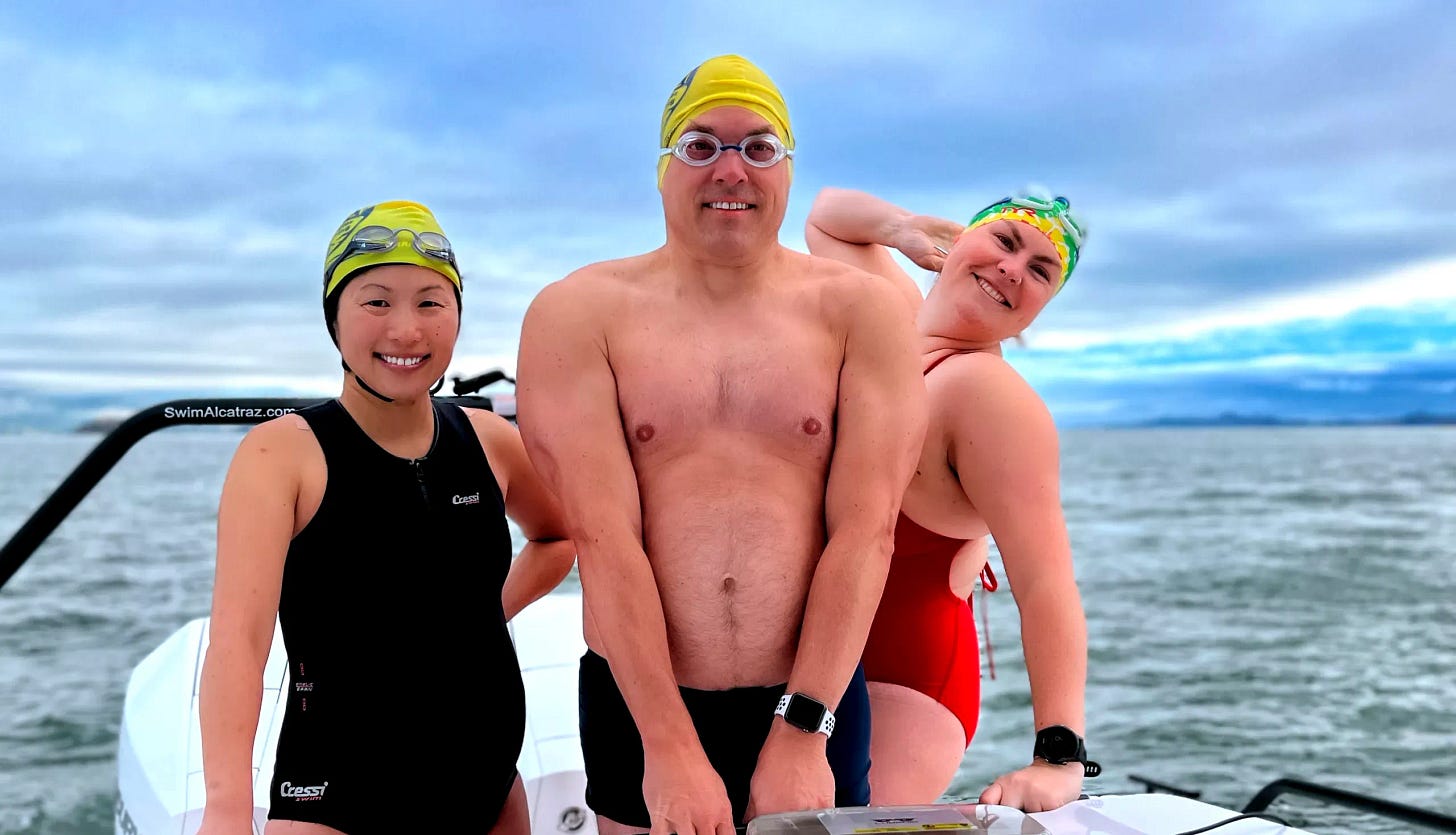 Three local San Francisco swimmers on a boat preparing for a Bridge-to-Bridge 10K swim, including one pregnant swimmer; cold water in background under cloudy skies.