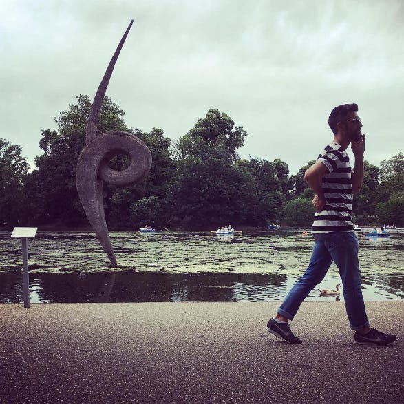 A pond in Victoria Park, East London, with people in rowing boats, trees in the background and a sculpture rising out of the water. A young man walks by the pond, talking into his cellphone. A pond in Victoria Park, East London, with people in rowing boats, trees in the background and a sculpture rising out of the water. A young man walks by the pond, talking into his cellphone.