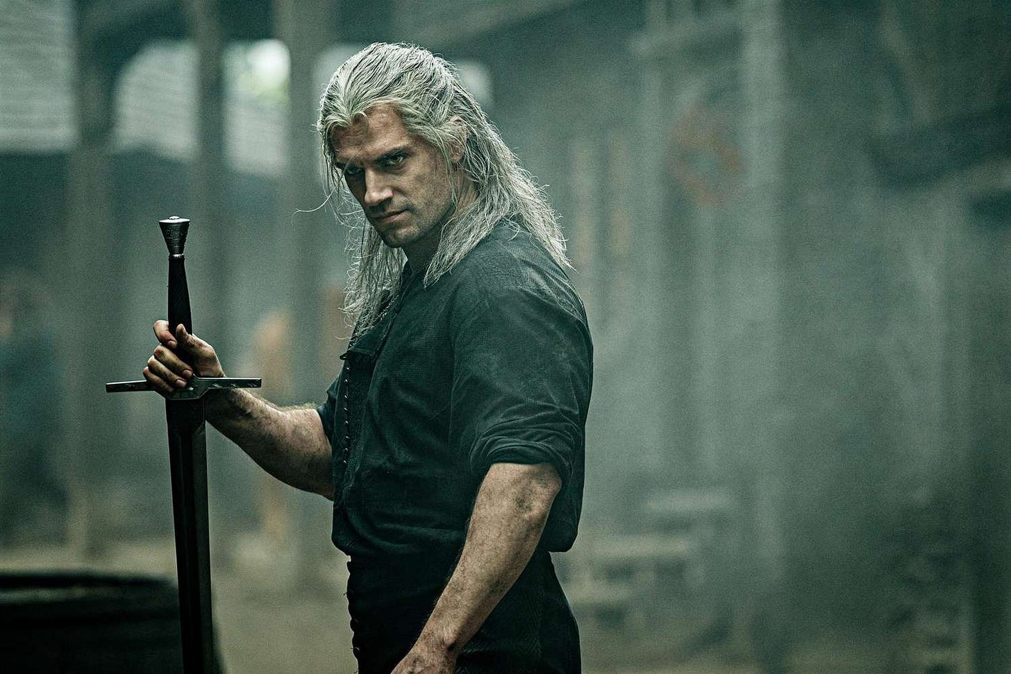 Henry Cavil as the Witcher, standing with sword out looking past camera at some bad guys. Buildings in background