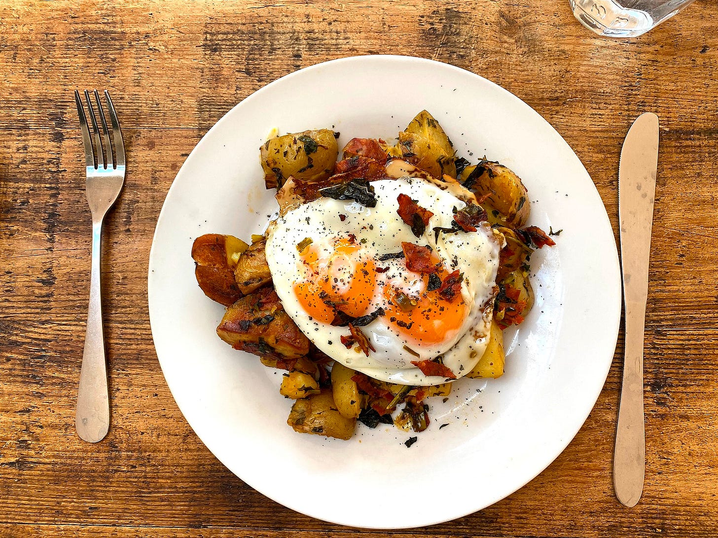 Image shows a round white plate on a wooden table. A knife and fork are placed either side. On the plate, there are potatoes and fried egg with two yolks.