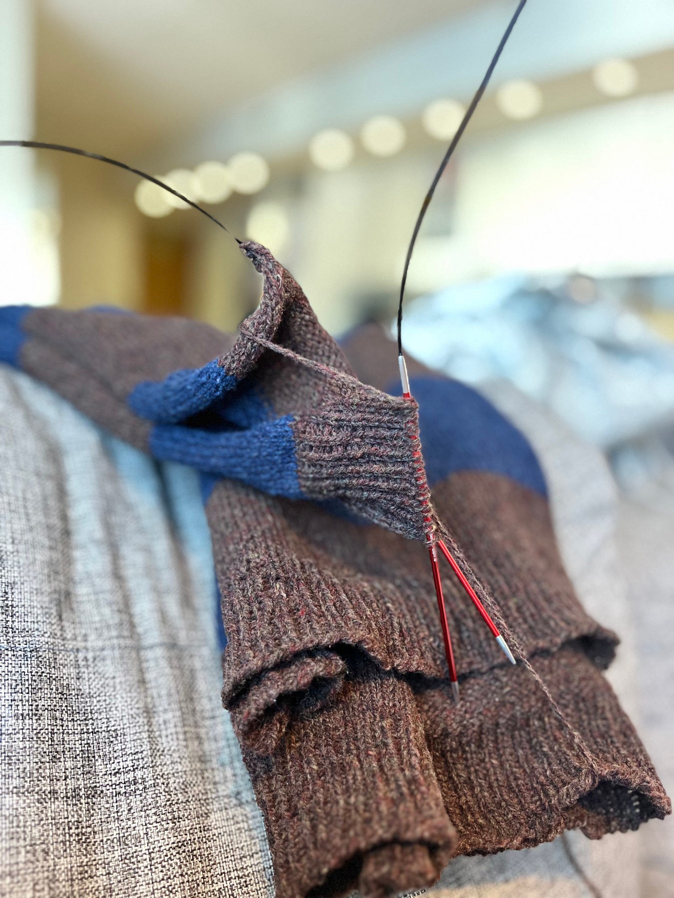 Close-up of a sweater being seamed, a tapestry needle working through the edges of chocolate and navy striped knitted pieces, yarn and circular needles visible.