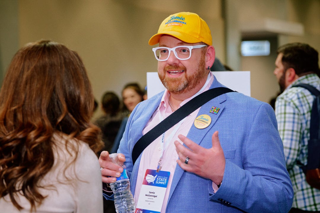 Jamie McConnigal in a yellow campaign baseball cap with white frame glasses and a blue blazer with a campaign button, speaking with one hand gesturing to someone off frame